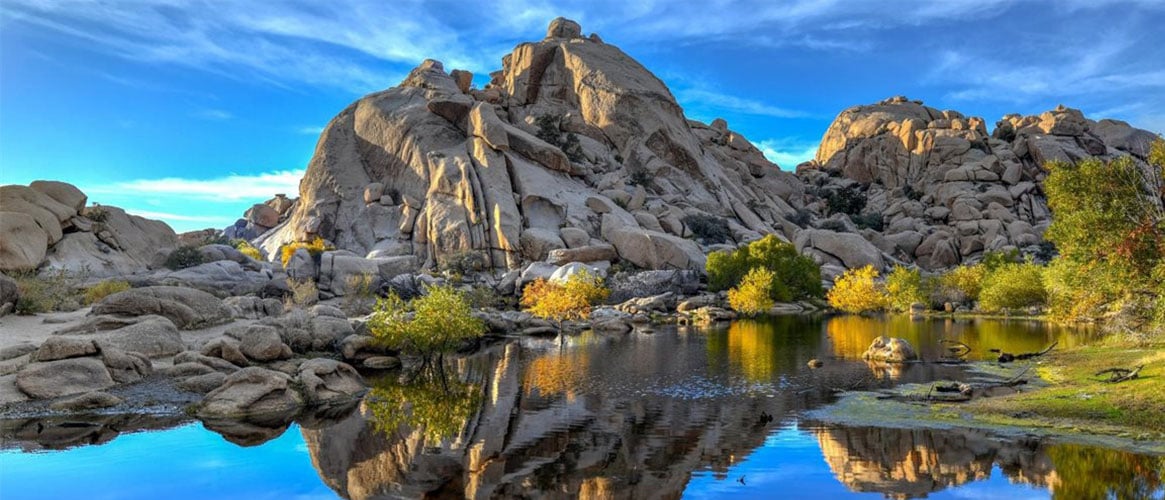 Massive desert boulder formations and golden autumn shrubs mirrored in a calm reflecting pool under a bright blue sky.